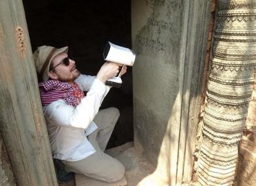 Giles Spence Morrow, crouched on the ground holding a white scanner at a site in Cambodia